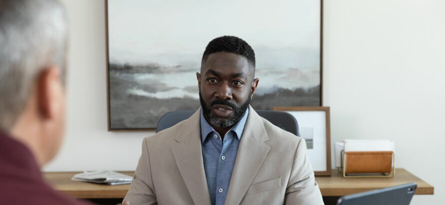 A man sits at a desk talking to someone across from him.