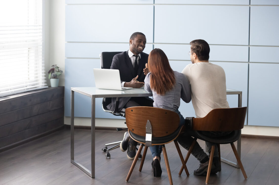 Happy smiling african american male realtor broker banker talking consulting young married couple. Full length millennial family spouse meeting real estate agent or financial advisor at office.