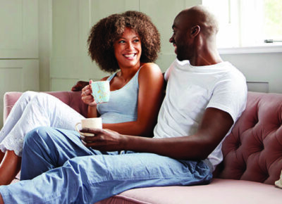 A young, smiling couple drinks coffee on a couch.