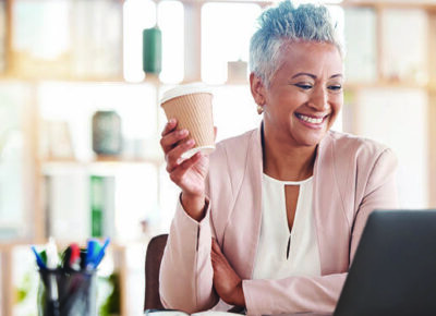 A woman holds a coffee and smiles at her laptop.
