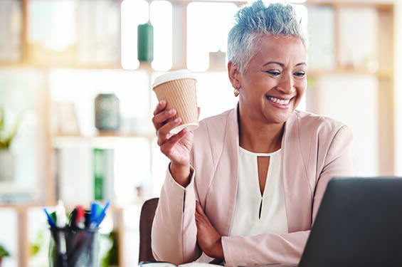 A woman holds a coffee and smiles at her laptop.